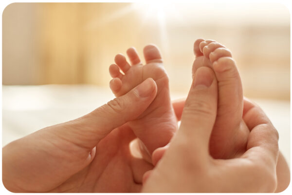 Mum making baby massage, mother massaging infant bare foot, preventive massage for newborn, mommy stroking the baby's feet with both hands on light background.